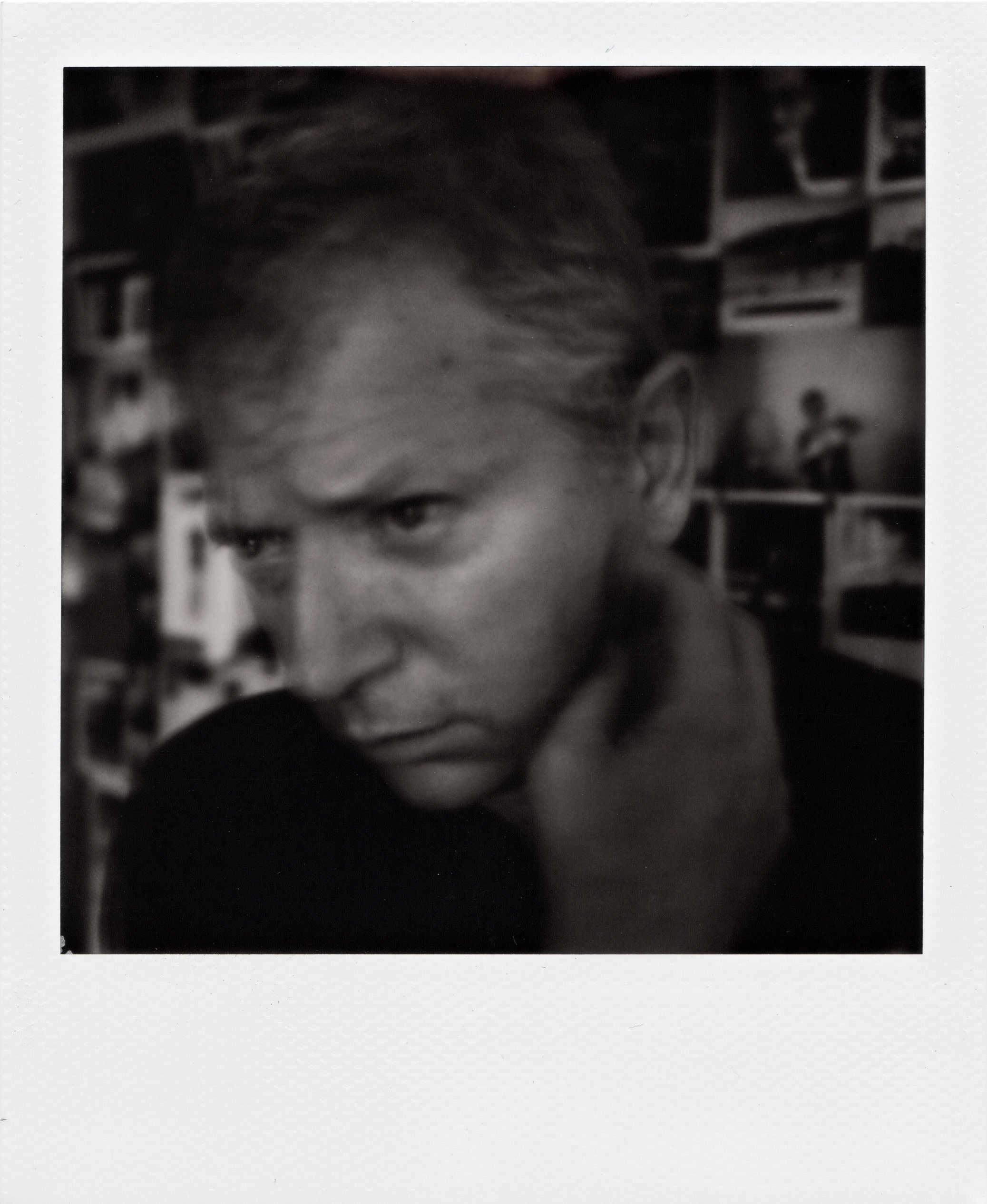 Ritratto in bianco e nero di un uomo adulto; volto in primo piano con espressione pensierosa, mano destra che tocca il collo sotto la mandibola, pelle chiaramente definita dalla luce laterale che crea ombre profonde sulle guance e attorno agli occhi; sfondo interno sfocato con dettagli indistinti.