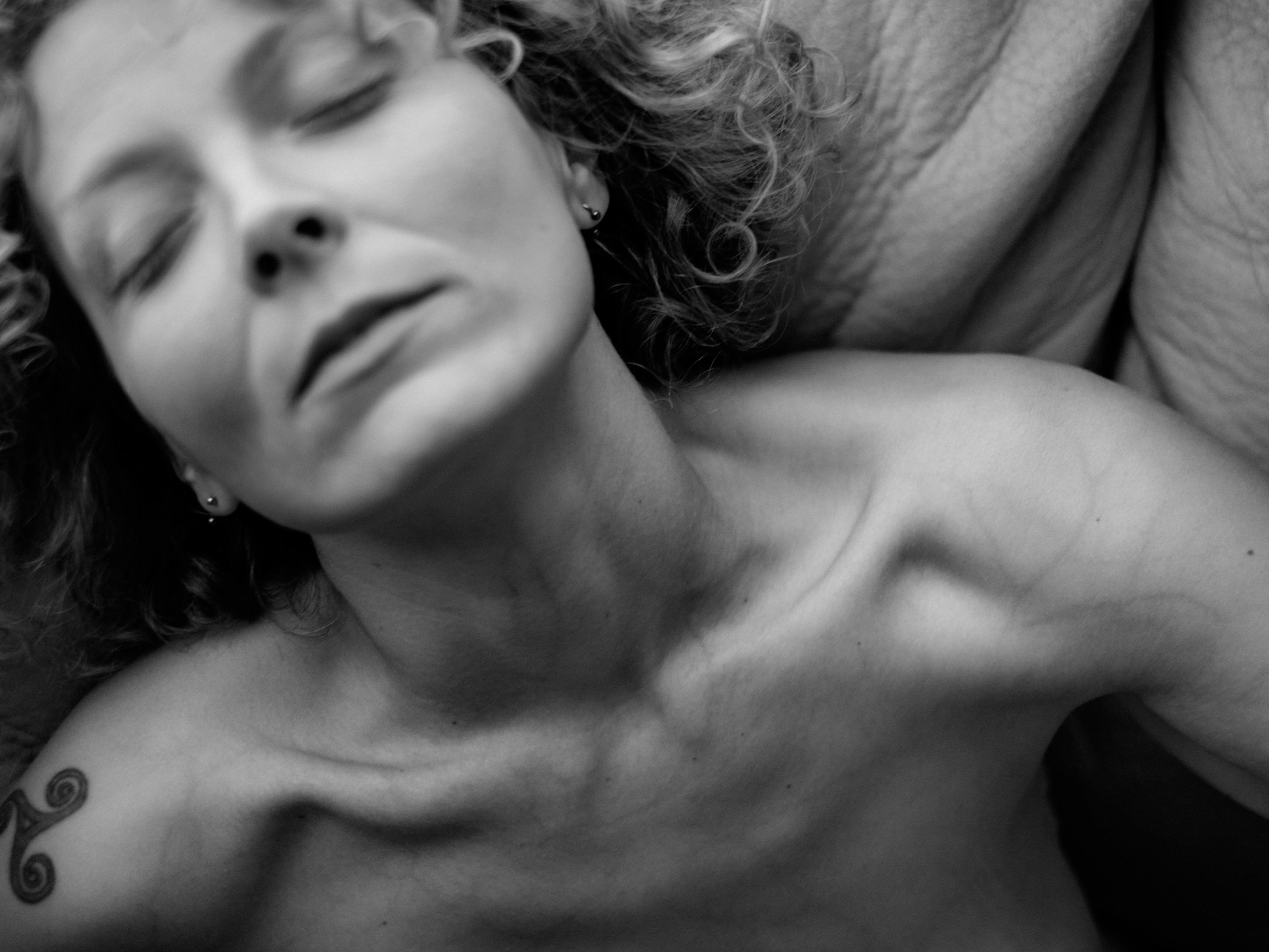 Primo piano in bianco e nero di una donna sdraiata o reclinata; testa inclinata indietro, occhi chiusi, collo e clavicole ben visibili, parte superiore del seno esposta; pelle con texture evidente e luce forte che crea ombre sugli zigomi e sotto il mento; sfondo scuro sfocato.