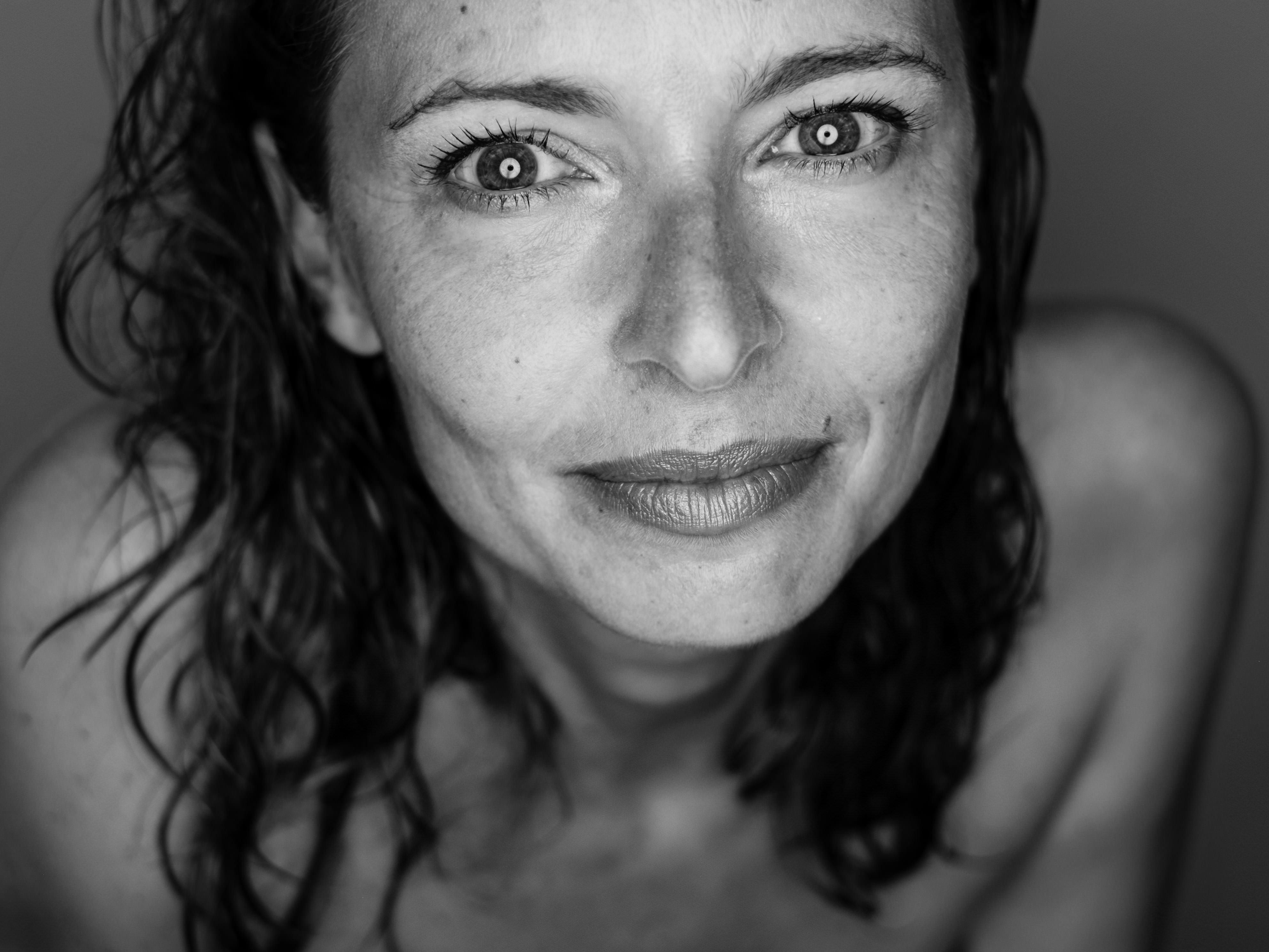 Primo piano in BN del volto con espressione serena; pelle chiara con lentiggini visibili, occhi aperti che guardano l'obiettivo, labbra chiuse e capelli mossi che cadono sulle spalle; luce frontale morbida che riduce le ombre; inquadratura mostra il viso fino alle spalle.
