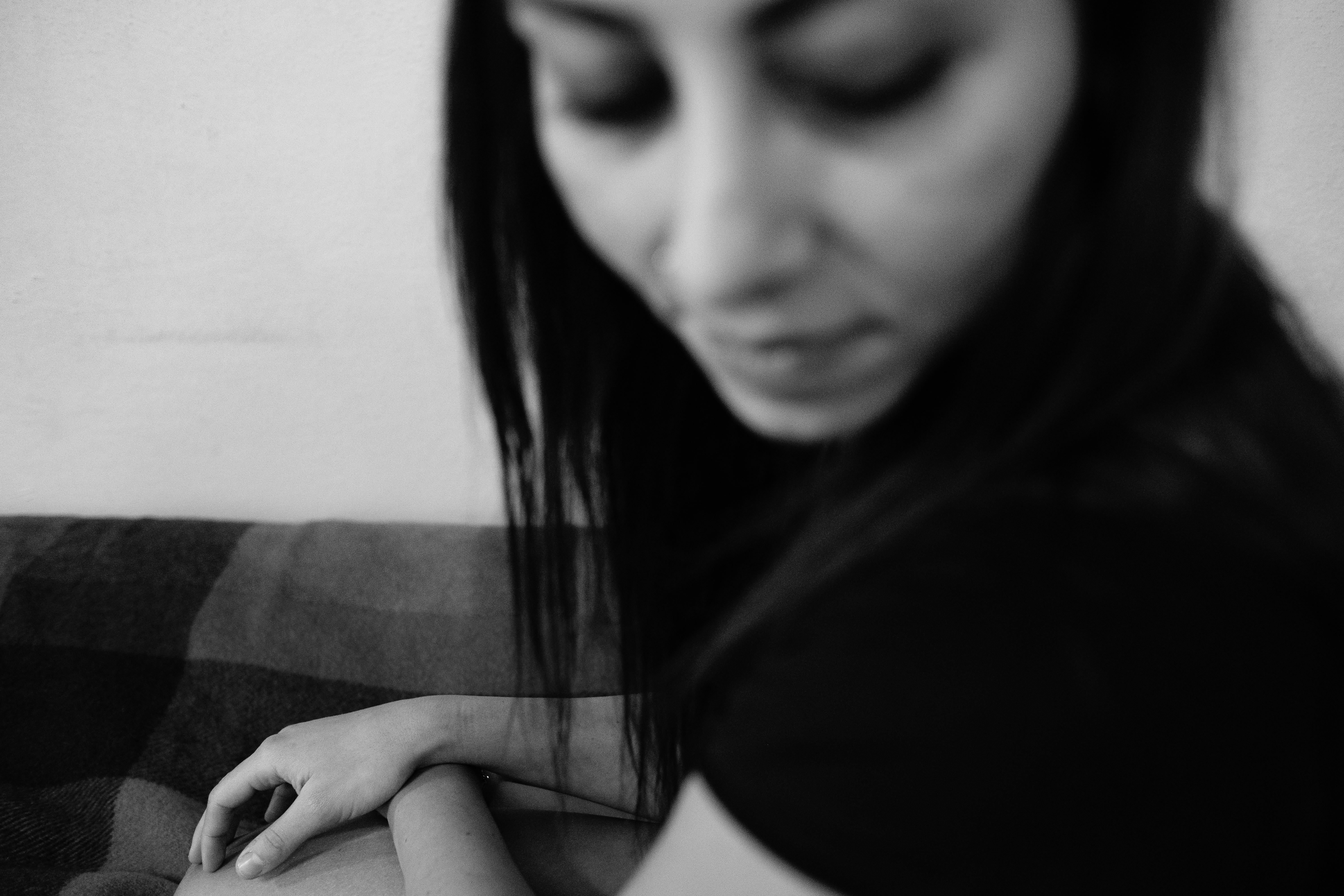 Ritratto in bianco e nero di una donna seduta su un divano con il busto leggermente inclinato in avanti; capelli lunghi scuri che cadono sul lato sinistro, espressione assorta con occhi rivolti verso il basso; inquadratura mostra il profilo del viso e parte del torso; luce morbida laterale che crea un alone luminoso sul viso.