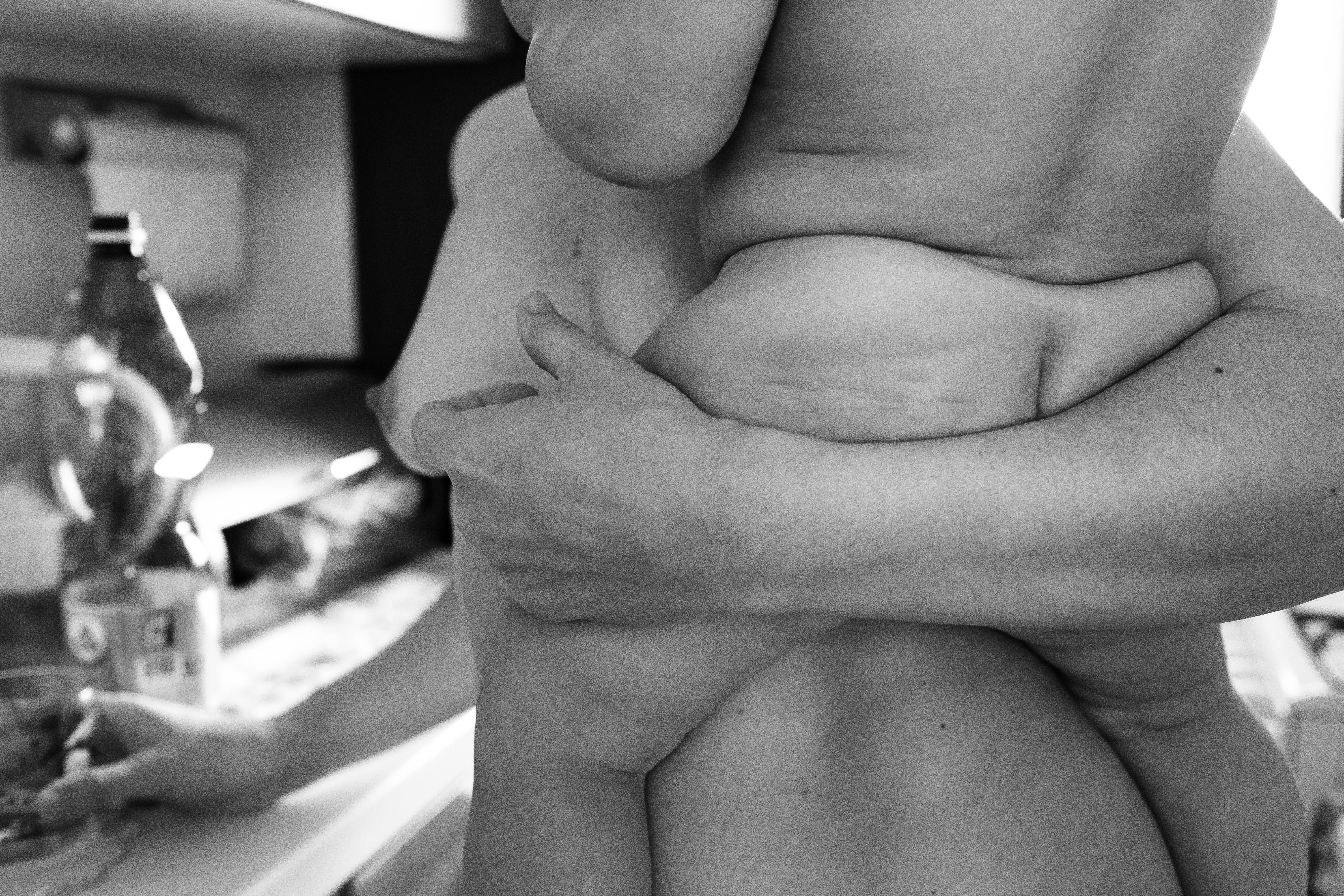 Scatto in bianco e nero che ritrae una madre e una figlia vicine nello stesso piano; inquadratura ravvicinata che mostra torsI e mani che si intrecciano mentre una tiene o culla un neonato vicino al petto dell'altra; pelle chiara, dettagli tattili delle mani e delle braccia, luce morbida che crea ombre delicate; sfondo domestico sfocato.