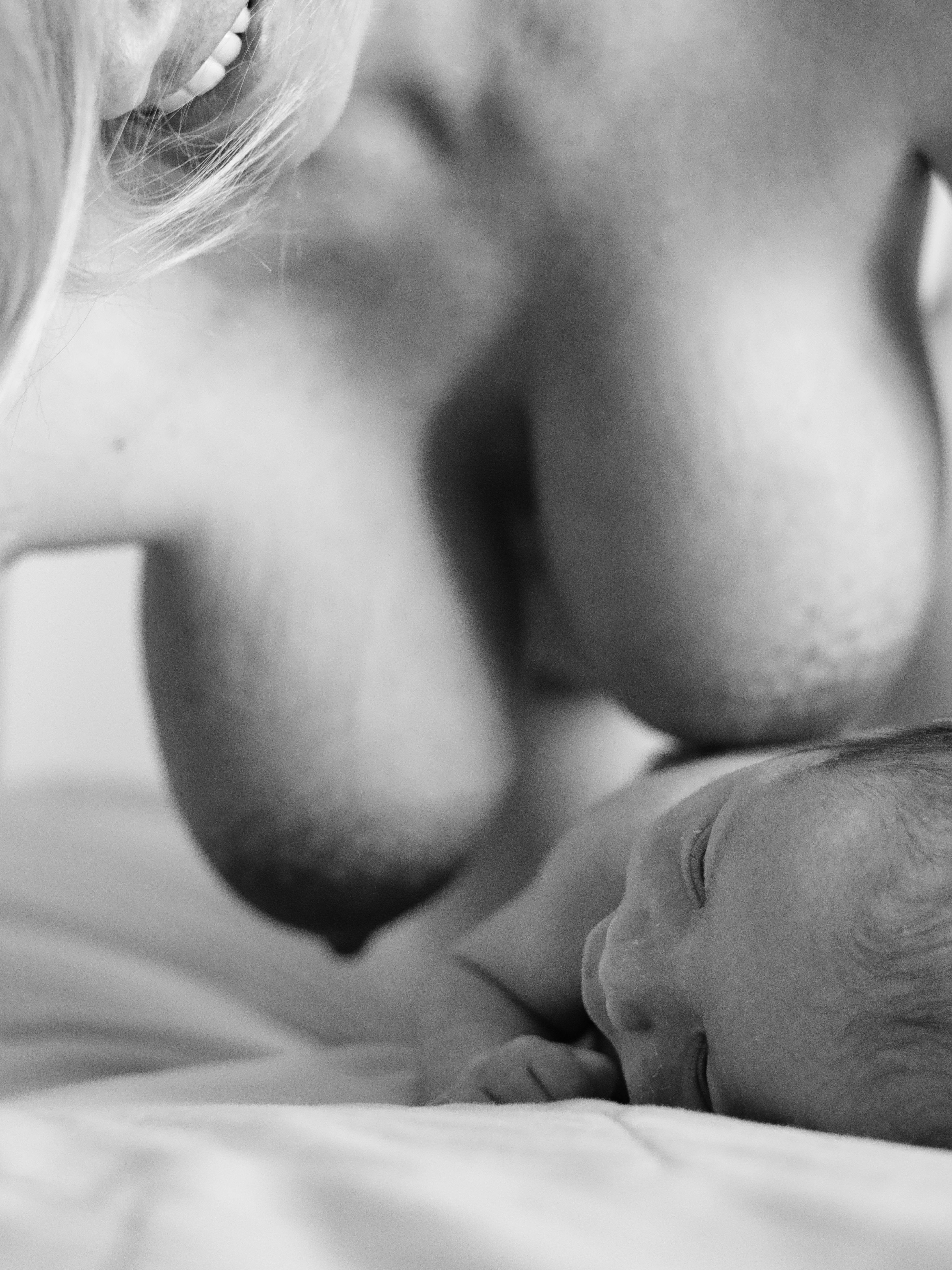 Fotografia in bianco e nero di una madre che tiene un neonato vicino al petto, inquadratura ravvicinata che mostra il profilo e il seno pesante della madre e la testa del bambino appoggiata al petto; pelle chiara, dettagli tattili delle mani e delle guance, luce morbida dall’alto che crea ombre delicate; sfondo neutro.