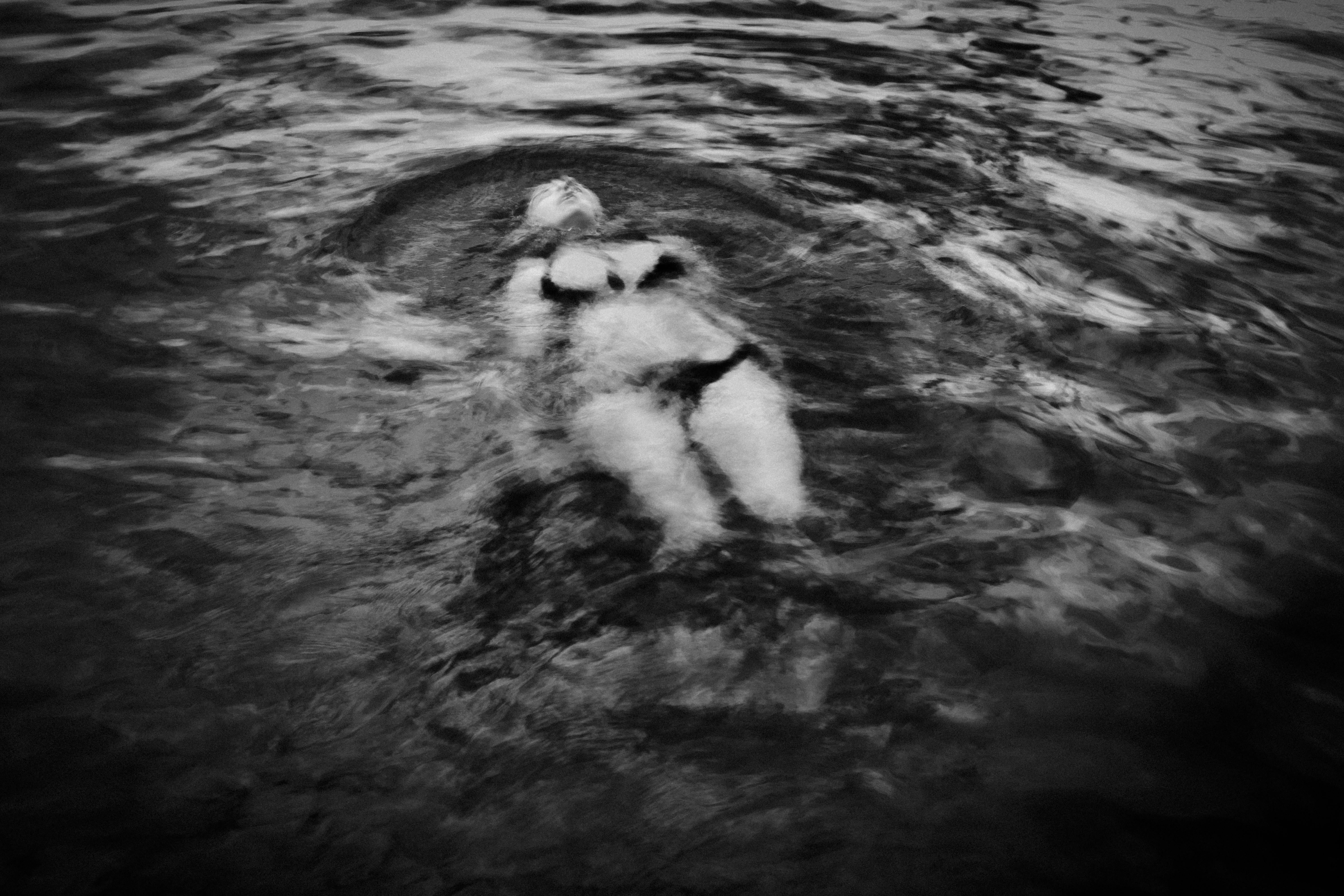 Fotografia in bianco e nero di una donna che galleggia sulla schiena in acqua; il corpo è immerso con viso e capo rivolti verso l'alto, occhi chiusi, braccia lungo i fianchi, increspature concentriche intorno al corpo; contrasto alto tra luce e acqua, dettagli delle bolle e delle superfici mosse.