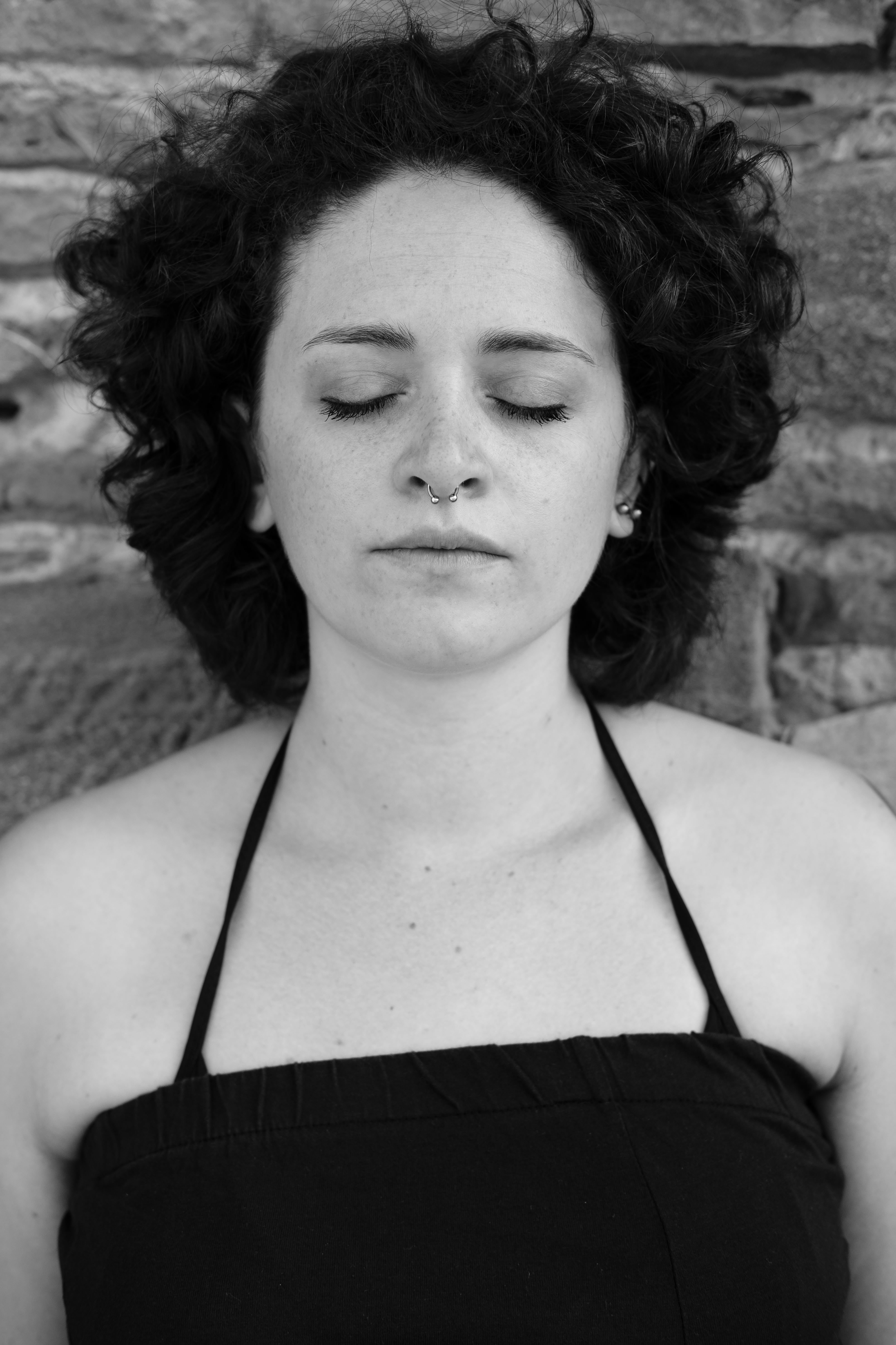 Ritratto in bianco e nero di una donna con capelli corti e ricci; occhi chiusi, volto rilassato, indossa un top nero con spalline sottili; luce frontale morbida, sfondo in pietra sfocato.