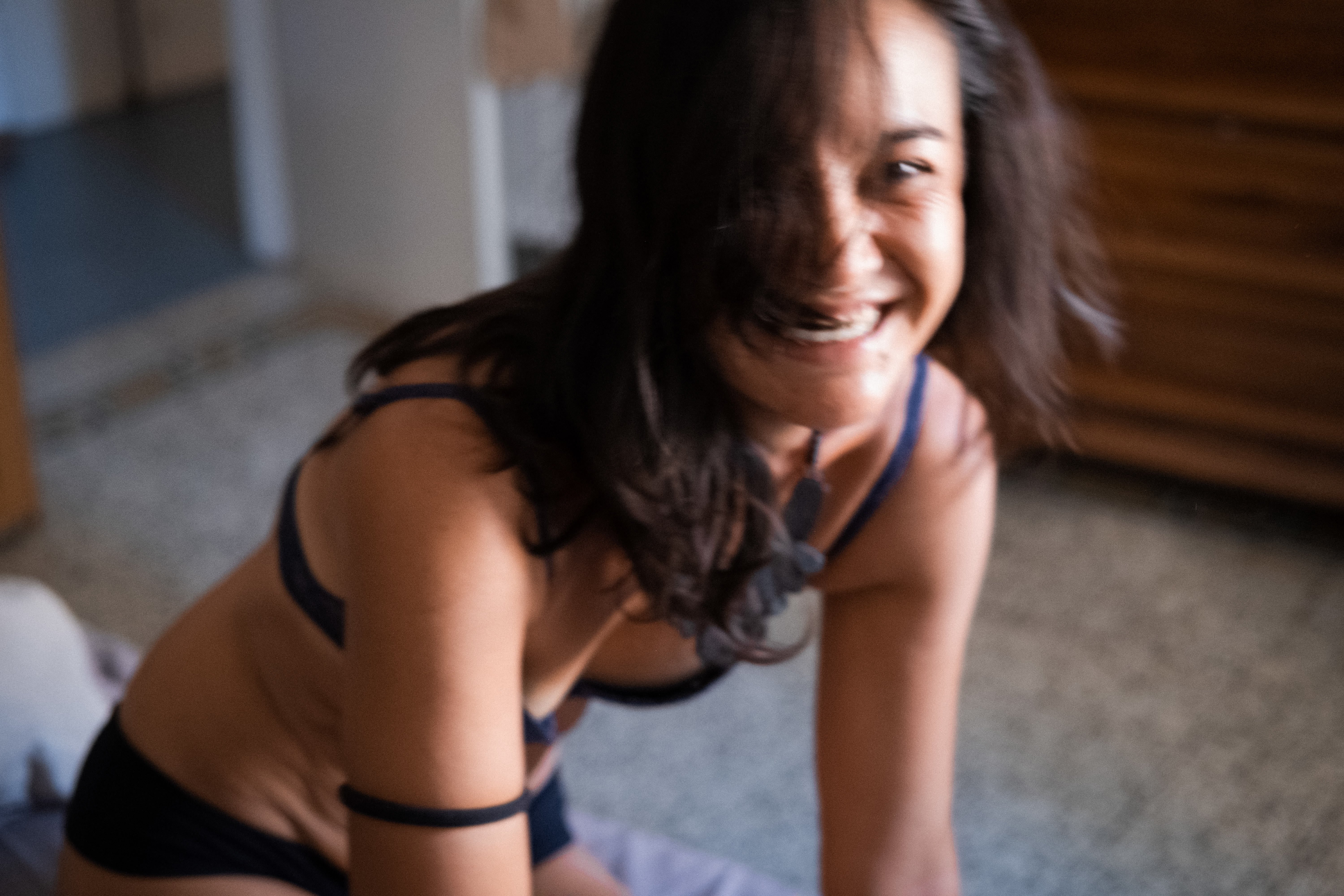 Ritratto a colori in interno: donna inginocchiata sul pavimento che si sporge in avanti sorridendo verso l’obiettivo. Capelli scuri sulle spalle; volto con occhi socchiusi, naso e bocca sorridente; collo e clavicole in vista; spalle e braccia tese a sostegno del corpo; seno coperto da un reggiseno nero con spalline sottili; addome nudo; anche e cosce piegate sotto di lei; slip neri. Luce naturale laterale che evidenzia pelle e linee del corpo; sfondo domestico sfocato con pavimento in graniglia e parete vetrata a destra.