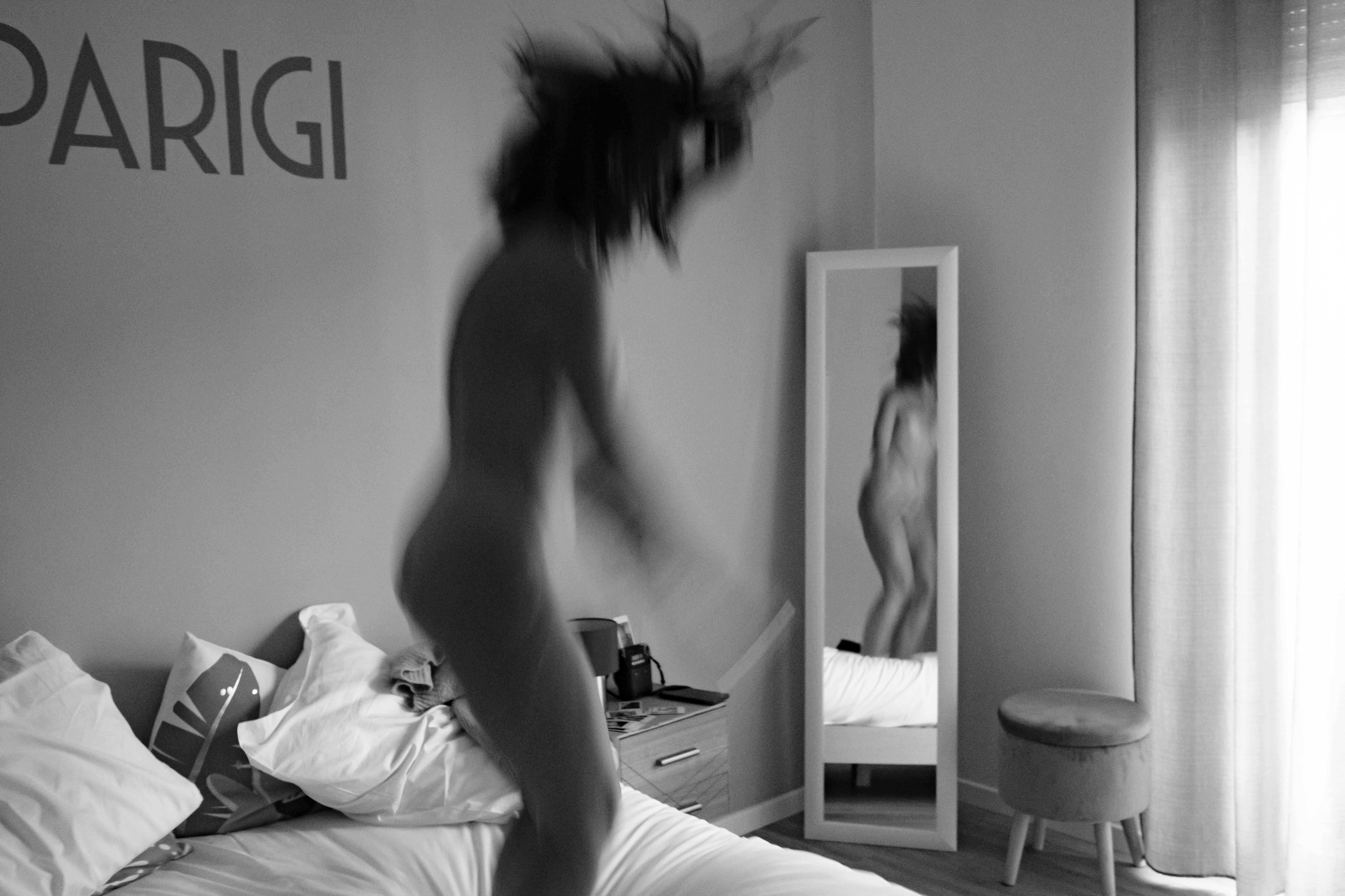 Scena in interno in bianco e nero con forte mosso: donna nuda che salta sopra un letto, corpo sfocato e slanciato; capelli che volano, braccia e gambe in movimento; seno nudo e ventre accennati dal mosso; grande scritta decorativa ‘PARIGI’ sulla parete; uno specchio sulla destra riflette parzialmente la sua nudità; finestra sullo sfondo.