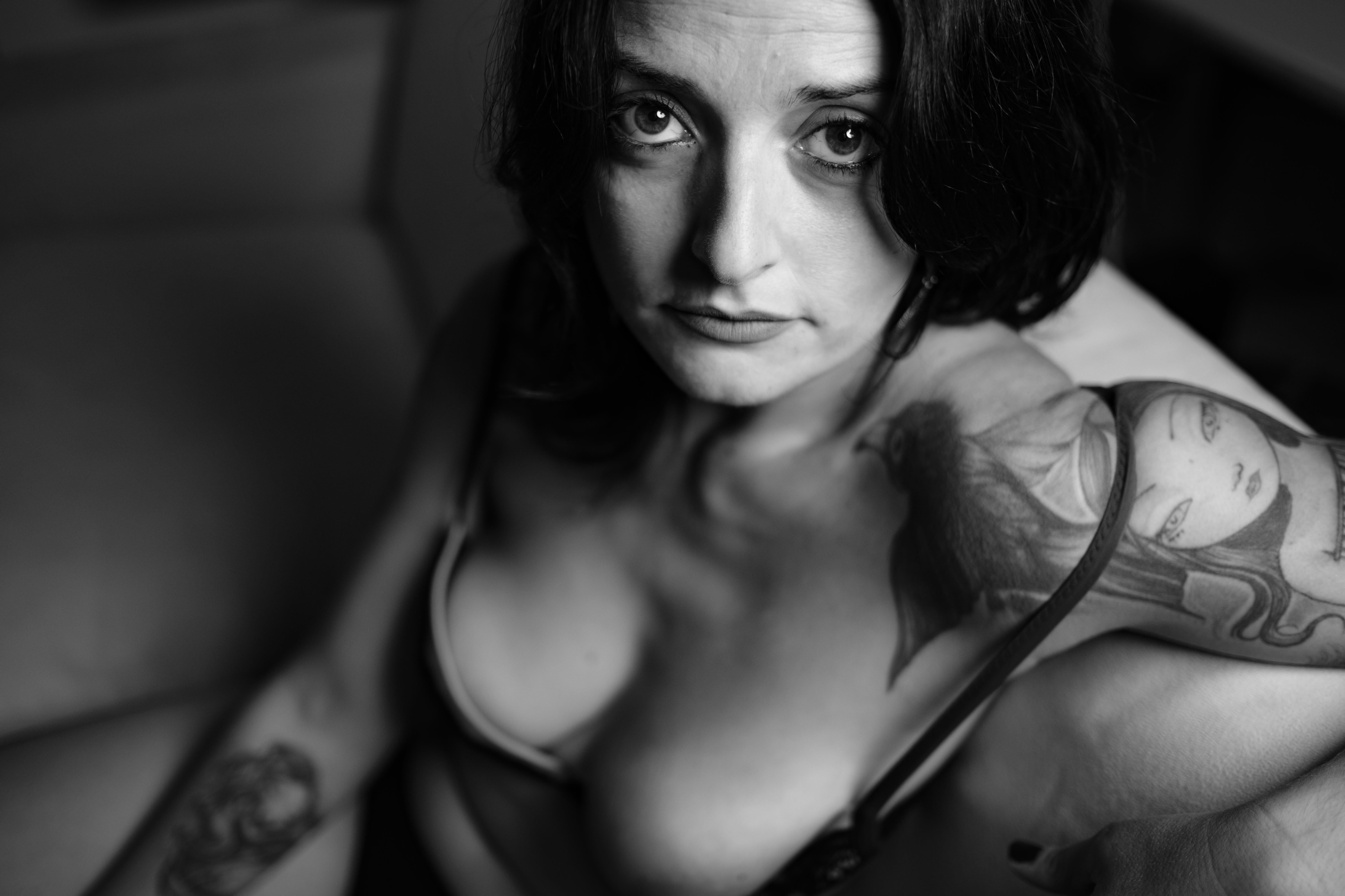 Ritratto ravvicinato in bianco e nero, mezza figura: donna seduta su un divano, sguardo diretto, capelli scuri al caschetto; braccia lungo i fianchi, tatuaggi su spalla e braccio; indossa un reggiseno con spalline sottili che copre il seno; luce morbida frontale che evidenzia occhi, naso e labbra.