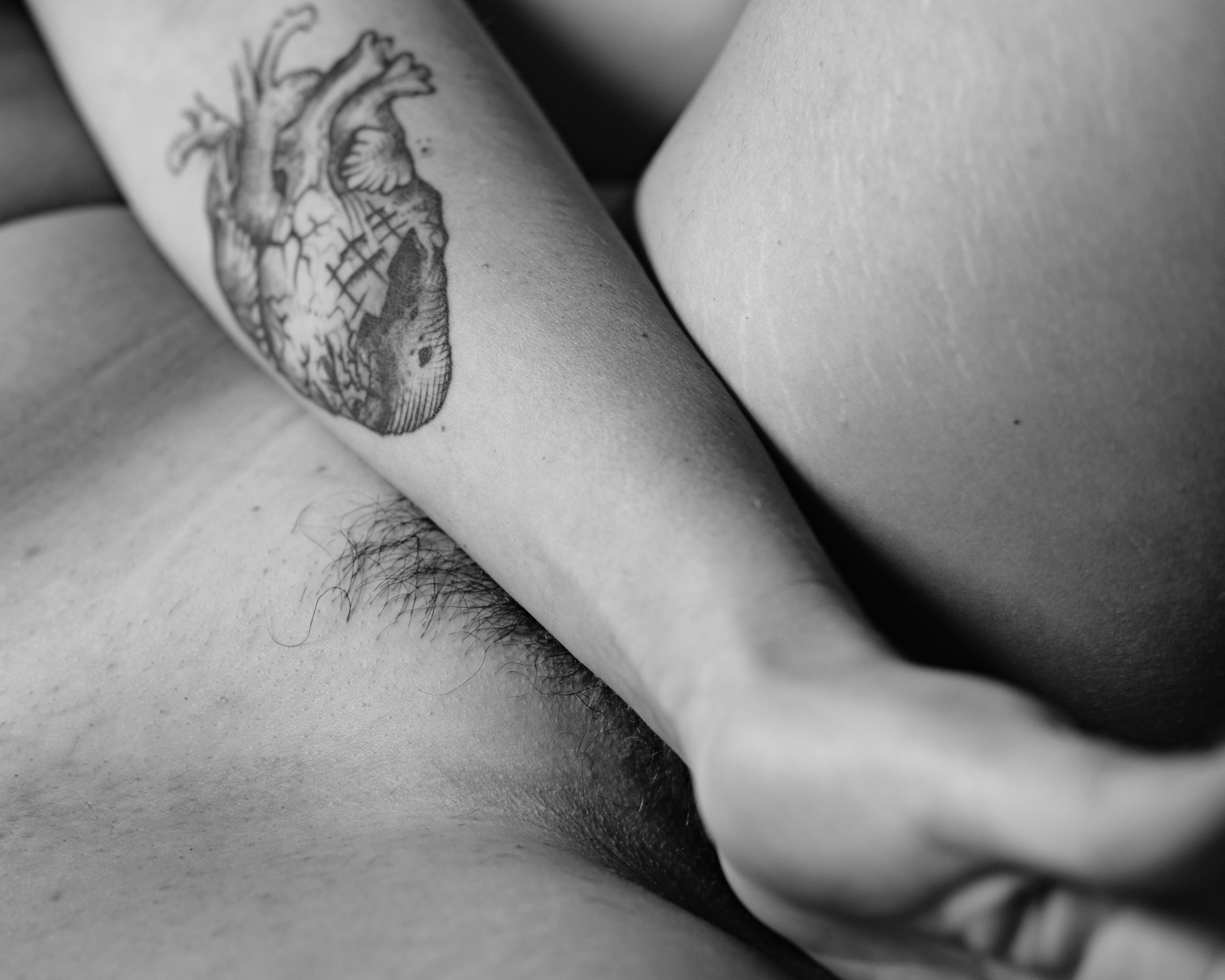 Dettaglio in bianco e nero della zona del bacino: cosce leggermente aperte, pube quasi completamente coperto dall’avambraccio che mostra un grande tatuaggio a forma di cuore; pelle illuminata da luce morbida; taglio stretto.