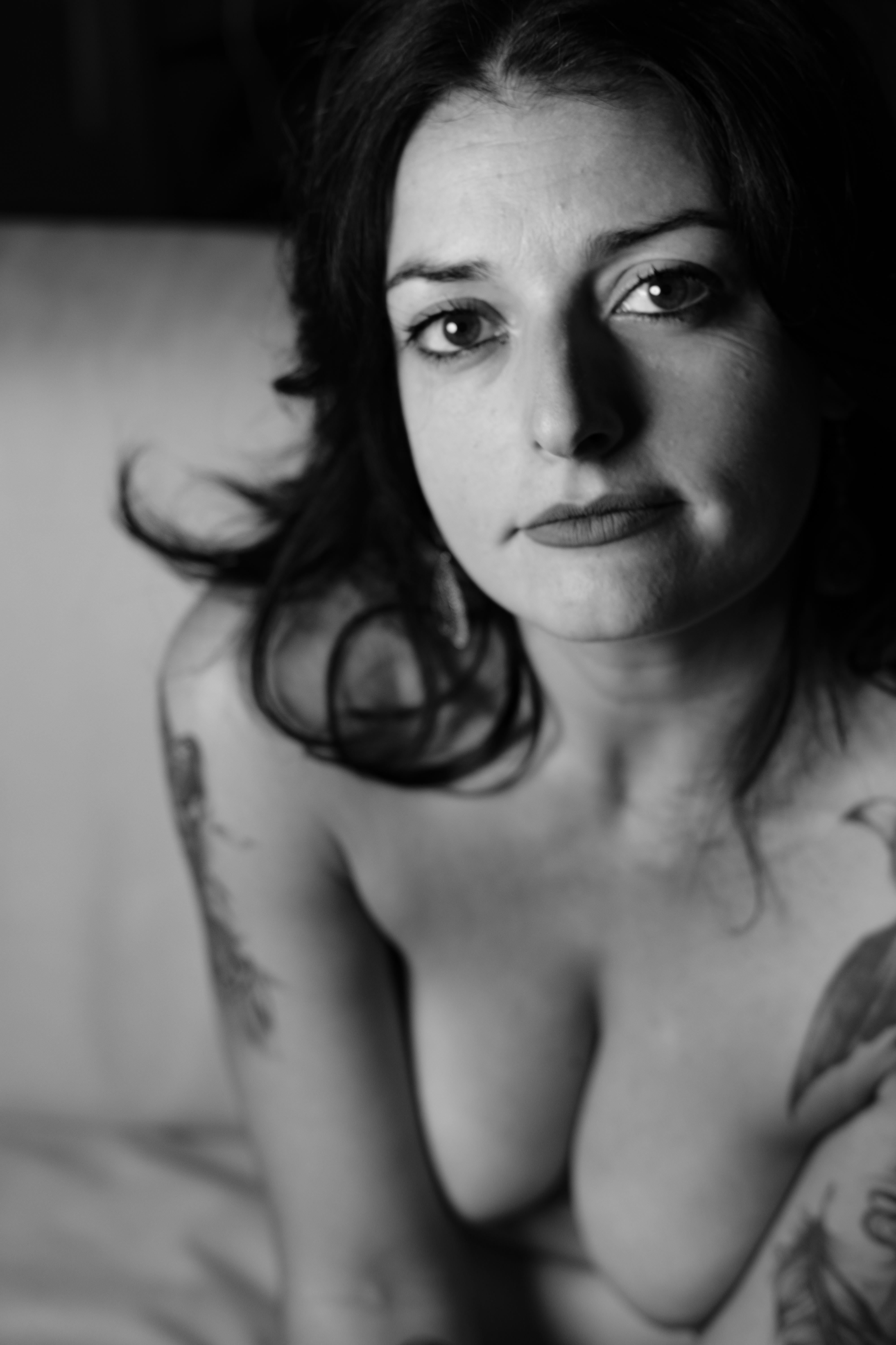 Ritratto ravvicinato in bianco e nero: donna seduta su un divano, spalle nude e seno in primissimo piano; volto frontale con sguardo diretto e leggero rossetto; capelli corti scuri; luce morbida che disegna clavicole, seno e lineamenti del viso.