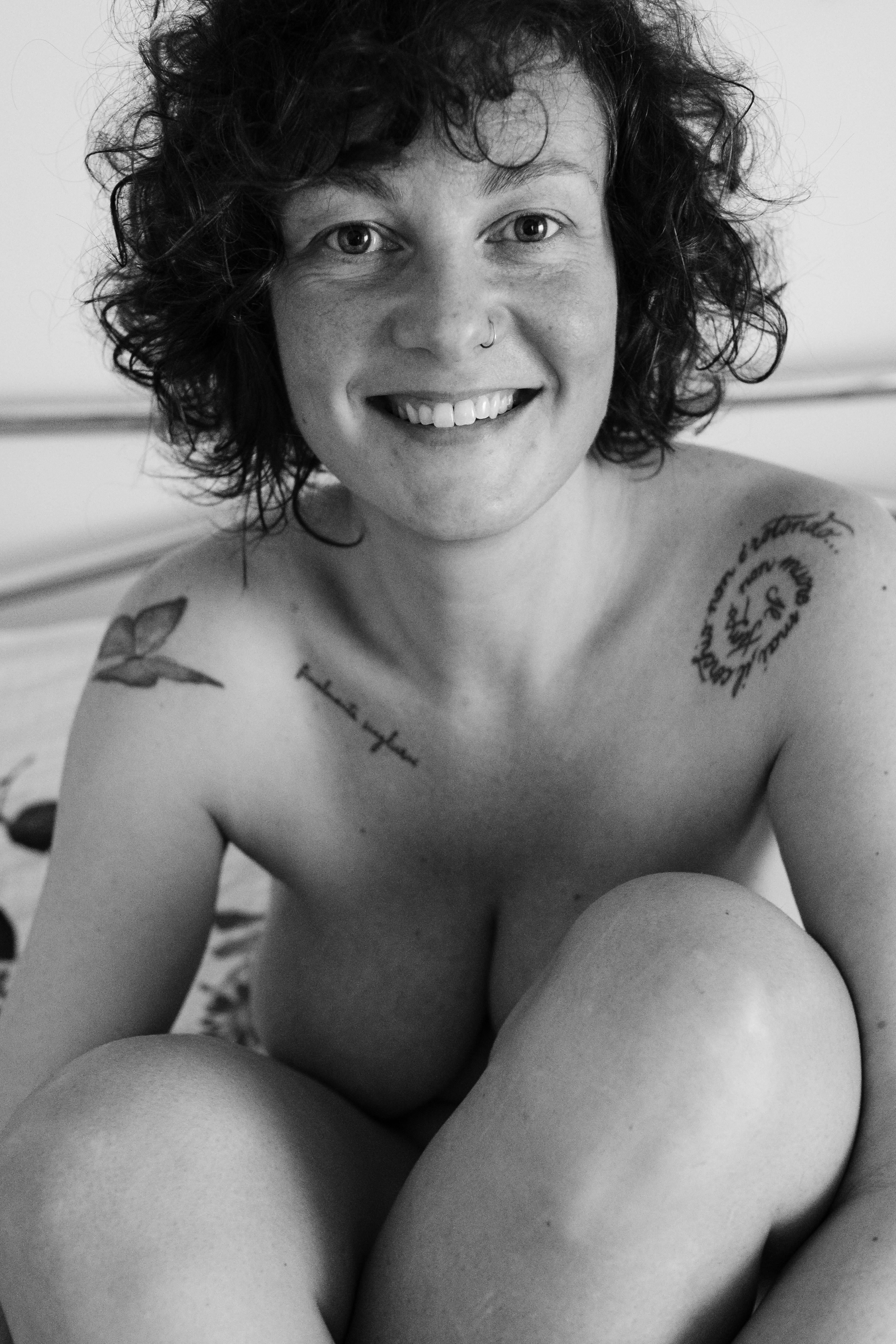 Ritratto in bianco e nero a mezzo busto: donna sorridente seduta su un letto, capelli ricci corti; sguardo diretto in camera; braccia piegate a cingere le ginocchia portate al petto; generoso seno nudo parzialmente coperto dalle braccia; tatuaggi visibili su spalla destra e braccio sinistro; luce morbida frontale.